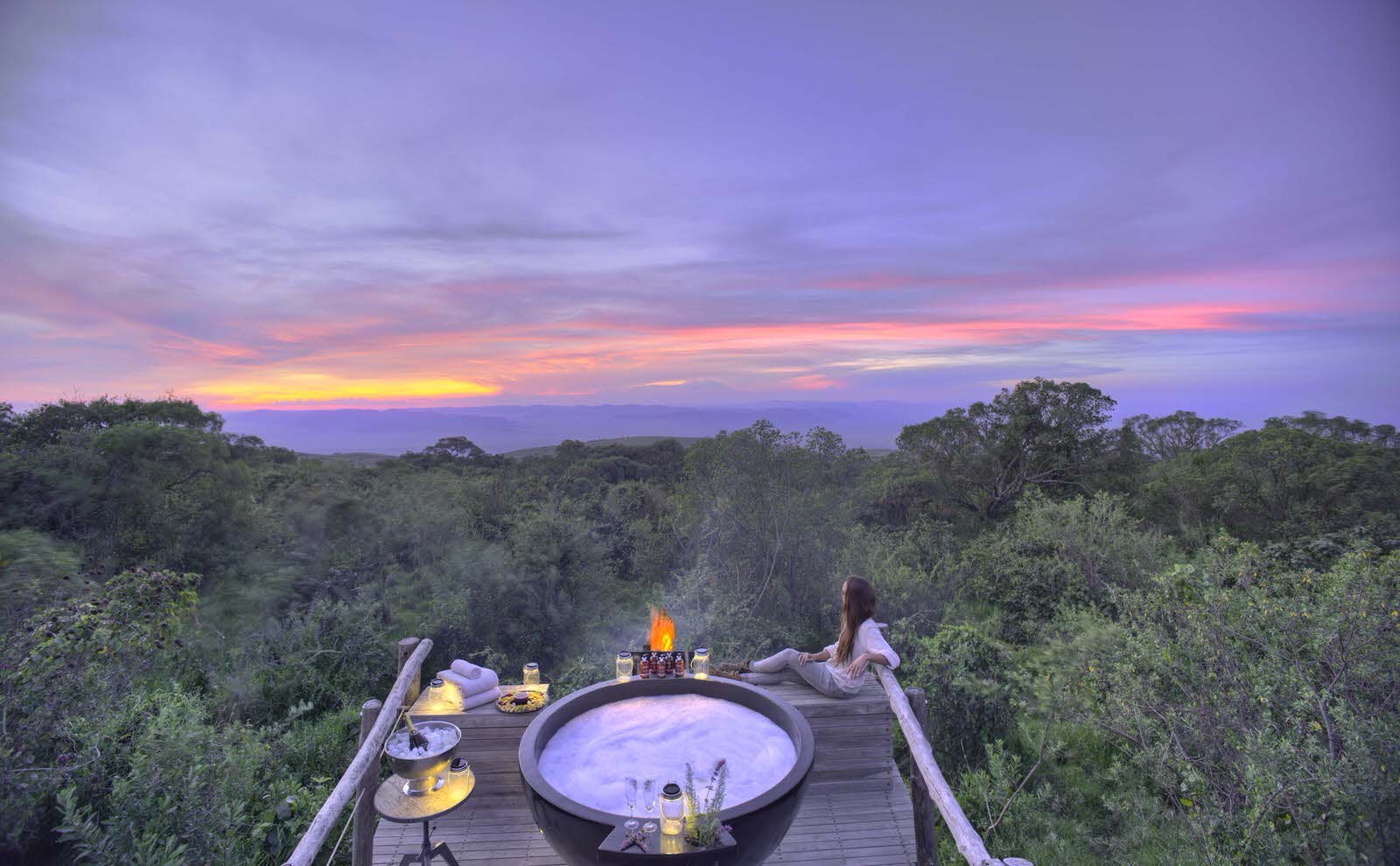 Features - luxurious-safari-accommodation-tanzania-d9775cfe870c A woman reclines on a wooden deck and looks out on the wilderness from a wooden deck at sundown. The deck has a hot tub, candles and small tables.