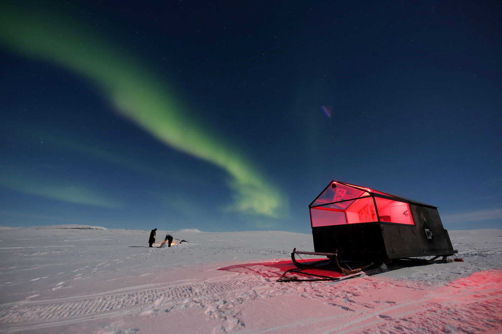 Features - camping-finland-wilderness-9a5911774b92 A small hut with a clear roof stands on large skis in the middle of a snowy, bare landscape with the Northern Lights in the sky and two people standing the background.