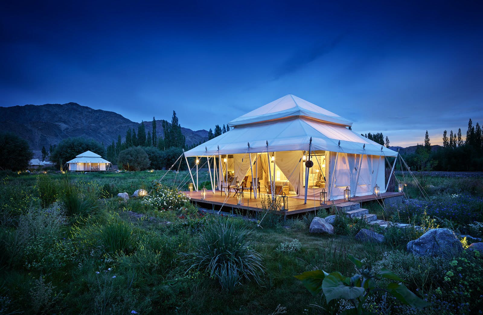 Features - remote-glamping-india-845e3317cf41 A square white tent with two-tired pyramidal roof glows with soft light in the dusk with the mountains of Ladakh in the background; the tent sits amongst lush vegetation, another similar tent is in the distance