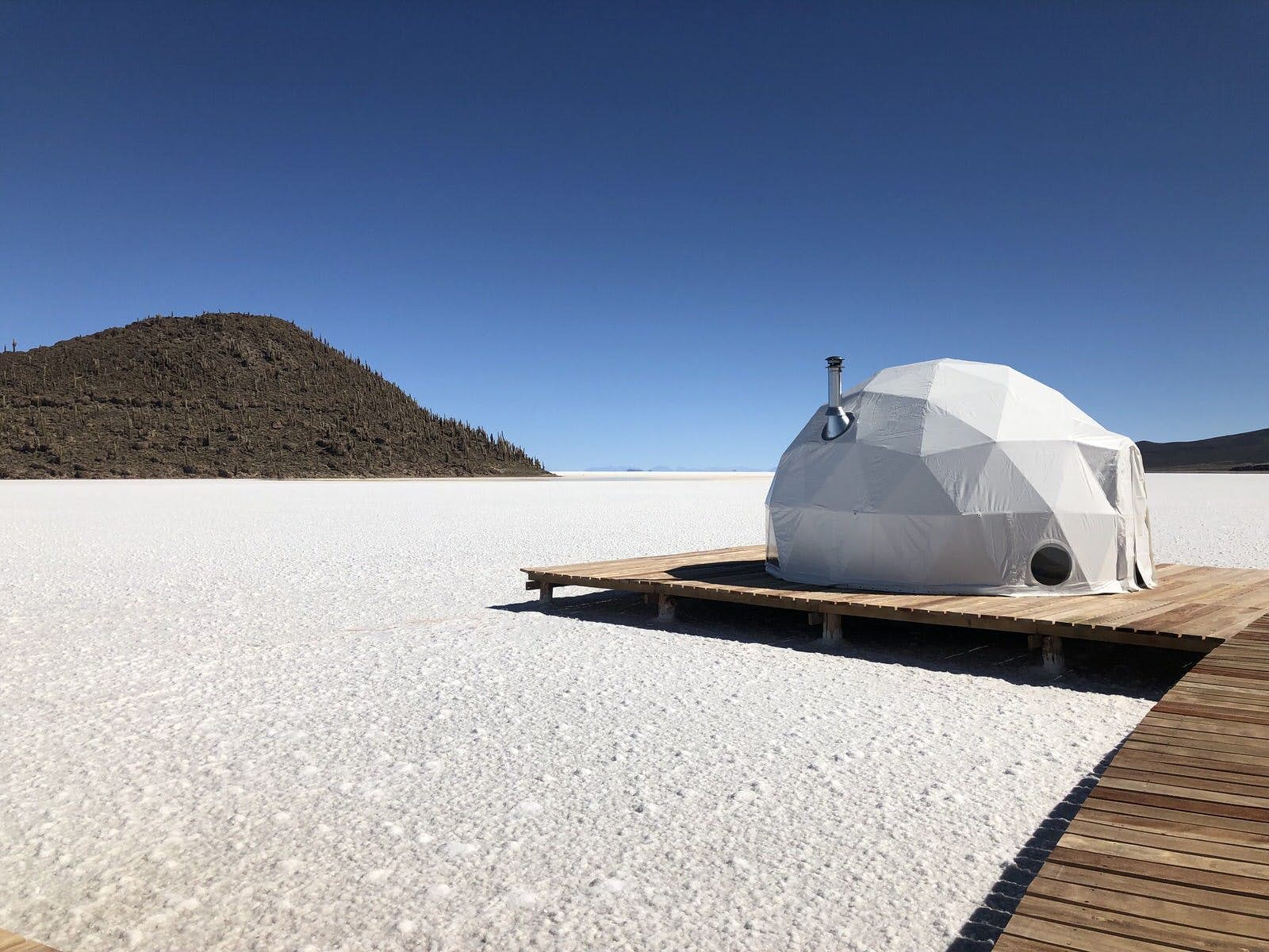 Features - stay-on-salt-flats-68232174c4bc A white geodesic domed tent with a silver chimney sits on a raised wooden platform, which seemingly hovers over white salt flats; in the semi-distance is a forested hill rising from the flats, much like an island